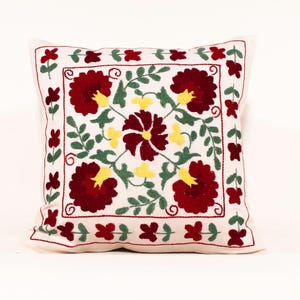 May include: Square white throw pillow with a detailed floral embroidery design. The embroidery features large burgundy flowers with yellow centers, green leaves, and small red flowers. The pillow is a decorative home accent.