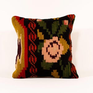 May include: Square decorative pillow with a floral design. The pillow features a black background with a large pink and green flower in the center. The edges have a geometric pattern in red, yellow, and burgundy.
