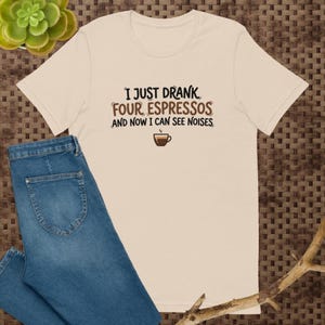 May include: A beige short-sleeve t-shirt featuring the black and brown text I JUST DRANK FOUR ESPRESSOS AND NOW I CAN SEE NOISES with a small brown coffee cup icon below the text, paired with blue denim jeans.