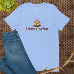 May include: A light blue t-shirt featuring a graphic of a smiling coffee cup with steam rising above it, surrounded by coffee beans. Below the cup, the text "Hello Coffee" is written in brown letters, with the word Coffee formed by beans.