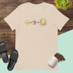 Può includere: Una t-shirt beige a maniche corte con una stampa grafica di un'equazione matematica. Il design mostra un'emoji gialla con faccia triste più una tazza di caffè marrone con un cuore uguale a un'emoji gialla di sole sorridente con raggi.
