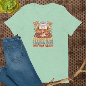 Puede incluir: Una camiseta verde claro con un gráfico de una taza de café sonriente abrazándose a sí misma, rodeada de granos de café y corazones. El texto dice COFFEE IS A LIQUID HUG FOR THE BRAIN en una tipografía colorida de estilo retro.