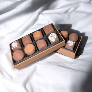 May include: Two open boxes of assorted chocolates. The chocolates are various shapes and sizes, with some decorated with white icing. The boxes are brown with black interiors, set against a white fabric background.