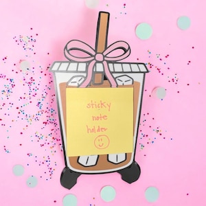 以下が含まれることがあります： ピンク色の背景に、バブルティーの形をした茶色と白の付箋ホルダー。ピンクのリボンと、"sticky note holder"とスマイリーフェイスが書かれた黄色の付箋が付いています。背景には紙吹雪と薄緑色の円が散りばめられています。