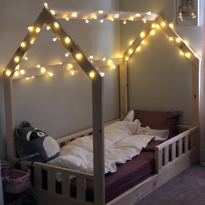 May include: A wooden bed frame with a house-shaped canopy, decorated with string lights. The bed has a brown mattress and a white comforter. A stuffed raccoon toy is on the bed.