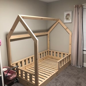 May include: A wooden toddler bed frame in the shape of a house with a slatted mattress and a railing around the sides. The bed is in a room with beige walls and carpet.
