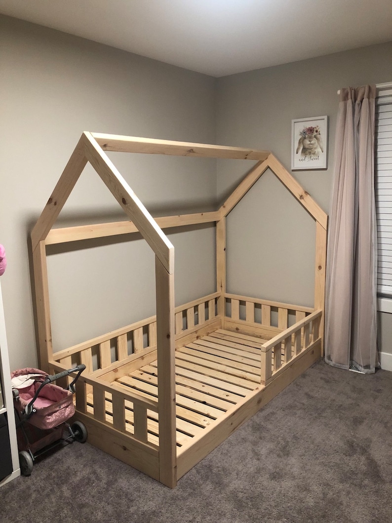 May include: A wooden toddler bed frame in the shape of a house with a slatted mattress and a railing around the sides. The bed is in a room with beige walls and carpet.