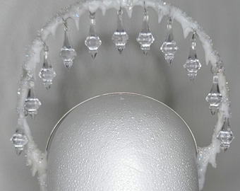 May include: A white halo crown with clear, teardrop-shaped crystal pendants. The crown is adorned with sparkling accents, creating a celestial or ethereal aesthetic. The design suggests a fantasy or costume theme.