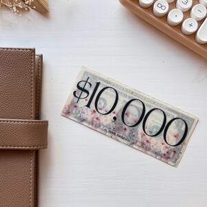 May include: A close-up of a $10,000 bill with a floral design. The bill is printed on a white background with pink flowers.