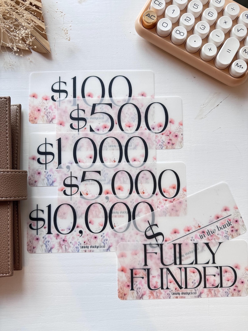 May include: Five transparent cards with a floral design and the text "$100", "$500", "$1,000", "$5,000", and "$10,000". A sixth transparent card with a floral design and the text "FULLY FUNDED in the bank".