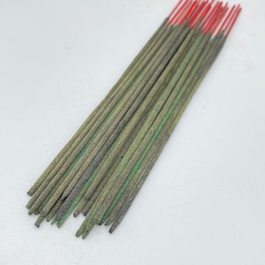 Puede incluir: Un conjunto de varillas de incienso con cuerpos verdes y marrones y puntas rojas. Las varillas de incienso están dispuestas en diagonal sobre una superficie blanca. Las puntas rojas contrastan con el verde y marrón apagados de las varillas.