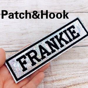 May include: A silver iridescent iron-on patch with black text that reads "FRANKIE" in a bold font. The patch has a black border.
