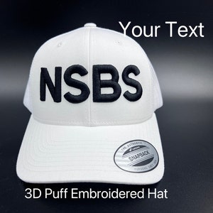May include: White trucker hat with a black 3D embroidered logo that says "NSBS". The hat has a mesh back and a snap closure. The hat is labeled "The Authentic Snapback Yupoong Since 1974".