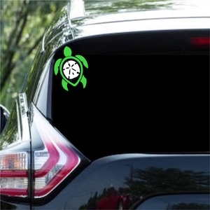 May include: Green turtle silhouette decal with a white sand dollar inside. The decal is on the back window of a black car.
