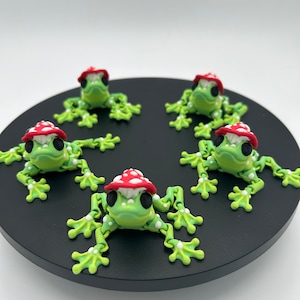 May include: Five green frog figurines with red and white mushroom hats. The frogs are posed in a circle on a black surface.