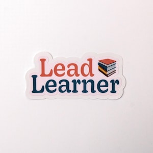 May include: A white sticker with the text "Lead Learner" in blue and orange. A stack of books is shown next to the text.