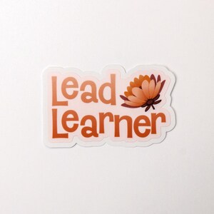 May include: A white sticker with the text "Lead Learner" in orange letters and a stylized orange flower graphic.
