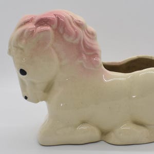 May include: A ceramic planter shaped like a white horse with pink accents. The horse has a mane and tail and is lying down with its head turned to the side. The planter has a hole in the top for drainage.