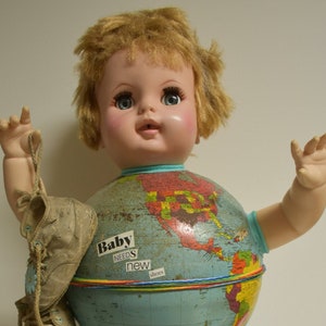 May include: A doll with blonde hair and blue eyes is dressed as a globe. The globe is blue and has a map of the world on it. The doll is holding a pair of brown shoes in one hand. The globe is sitting on a metal stand. The text on the globe reads "Baby Needs New Shoes".