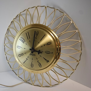 May include: A gold-toned Sessions clock with a round face and a decorative, openwork frame. The clock face features black numerals and hands, with the Sessions logo. The frame has a sunburst design. The clock is plugged into an electrical outlet.