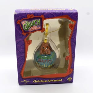 May include: A Christmas ornament featuring the Grinch character inside a clear, teardrop-shaped bauble. The ornament is in a purple box with the text "How the Grinch Stole Christmas!" and "Christmas Ornament". The ornament has a gold string for hanging.