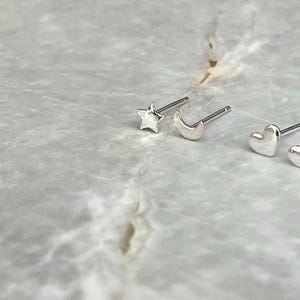 May include: A collection of three pairs of small, silver-colored stud earrings. One pair features star shapes, another crescent moons, and the last hearts. The earrings are displayed on a light gray, marbled surface.