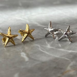 May include: Two pairs of starfish earrings. One pair is gold-coloured, and the other is silver-coloured. The earrings are small, with a three-dimensional design. The earrings are displayed on a marble surface.
