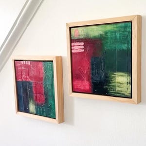 May include: Two square abstract paintings in light wood frames. The canvases feature textured layers of deep red, forest green, navy blue, and pale yellow paint with small pink accents. The artwork displays a grid-like composition with visible brushstrokes and raised paint application throughout the surfaces.