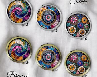 May include: A collection of round compact mirrors with mosaic-style designs. The mirrors are available in silver and bronze finishes, each featuring unique colorful patterns. The designs include spiral, geometric, and floral motifs, with the letters A, B, and C labeling each mirror.