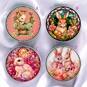Puede incluir: Cuatro objetos decorativos redondos con diseños temáticos de Pascua. Uno presenta un conejito en una cesta de flores, otro un conejito estilo vidriera, y otros dos muestran conejitos rodeados de flores. Cada uno tiene un borde de metal plateado.