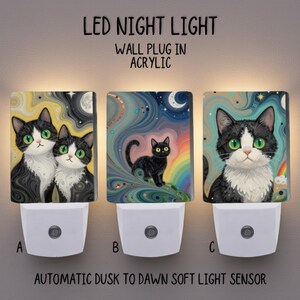Puede incluir: Tres luces nocturnas LED con ilustraciones de gatos. Cada una tiene una base blanca y un frente de acrílico. La primera presenta dos gatos con ojos verdes sobre un fondo amarillo y negro. La segunda muestra un gato negro sobre un arcoíris. La tercera tiene un gato con ojos verdes.
