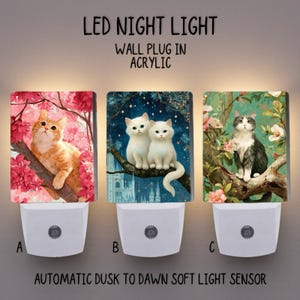 May include: Three LED night lights with cat illustrations. Each light has a white base and an acrylic panel. The panels feature a ginger cat in a cherry blossom tree, two white cats on a branch, and a grey and white cat in a floral setting. Text reads: LED NIGHT LIGHT, WALL PLUG IN ACRYLIC, and AUTOMATIC DUSK TO DAWN SOFT LIGHT SENSOR.