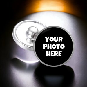 May include: A silver-colored snap button with a black, round face. The face has the white text "YOUR PHOTO HERE". The button is designed to attach to fabric or other materials.