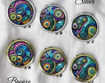 May include: A collection of six round compact mirrors with colorful, abstract designs. Three mirrors have silver frames, and three have bronze frames. Each mirror features a unique mosaic-like pattern in shades of blue, green, purple, and yellow.