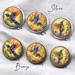 Könnte beinhalten: Silberne und bronzefarbene Pillendosen mit bunten Kolibri-Designs. Jede Dose zeigt eine farbenfrohe Kolibri-Illustration vor einem gelben floralen Hintergrund. Die Dosen sind rund und haben einen Klappdeckel. Die Wörter "Silver" und "Bronze" sind auf dem Bild geschrieben.