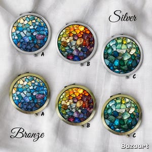 Könnte beinhalten: Eine Sammlung runder Kompaktspiegel mit Mosaik-Designs. Die Spiegel sind in Silber- und Bronze-Ausführungen erhältlich, jeweils mit farbenfrohen, unregelmäßig geformten Teilen in Blau-, Grün-, Orange- und Lilatönen. Die Wörter "Silver" und "Bronze" sind auf dem Bild geschrieben.