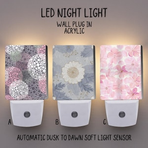May include: Three LED night lights with acrylic designs. The designs include a pattern of white and pink circles, a floral pattern with a white flower, and a pattern of pink flowers. The image text reads "LED NIGHT LIGHT" and "AUTOMATIC DUSK TO DAWN SOFT LIGHT SENSOR."