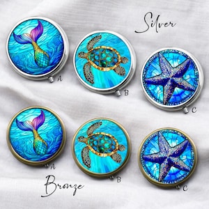 Puede incluir: Pastilleros redondos plateados y bronce con diseños de vida marina. Los diseños incluyen una cola de sirena, una tortuga marina y una estrella de mar. Los estuches tienen un fondo azul y turquesa.