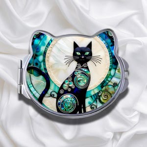 May include: A cat-shaped compact mirror with a black cat design. The cat has green eyes and is decorated with blue and green swirls. The mirror is framed in silver and set against a white background.
