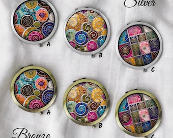 May include: Six round compact mirrors with colorful mosaic designs. Three have silver frames, and three have bronze frames. The designs feature swirls and geometric patterns in various colors, including blue, orange, and pink. The word "Silver" and "Bronze" are written above the mirrors.
