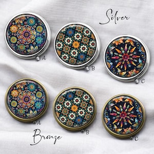 May include: Round pill cases with colorful floral designs. The cases are available in silver and bronze finishes. Each case features a different intricate pattern in shades of blue, orange, and purple. The cases are labeled A, B, and C.