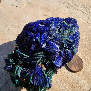 May include: Azurite and malachite crystal cluster with a blue and green color pattern. The crystal is sitting on a light brown surface next to a US penny.