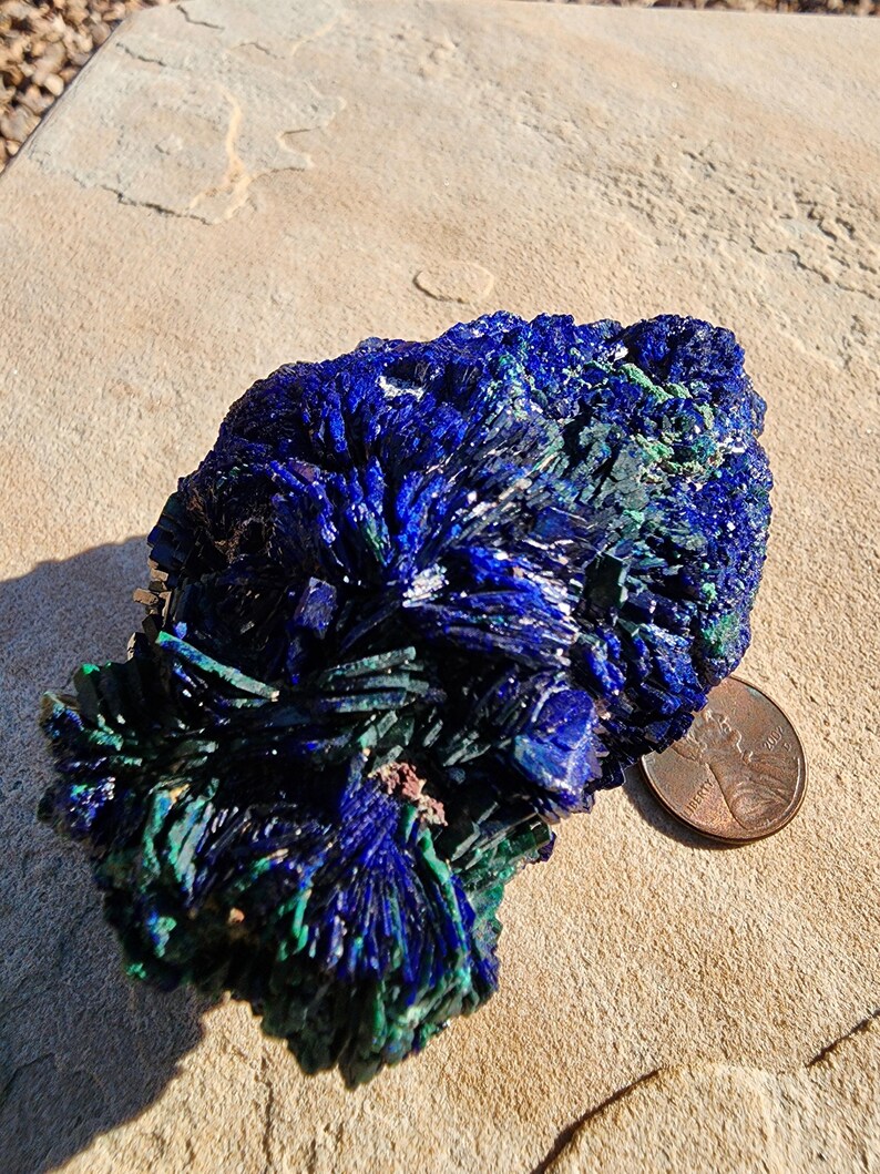 May include: Azurite and malachite crystal cluster with a blue and green color pattern. The crystal is sitting on a light brown surface next to a US penny.