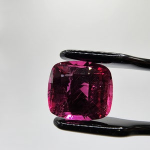 K&ouml;nnte beinhalten: Ein leuchtend rosa Edelstein mit Kissenform, gehalten von einer schwarzen Metallzange. Der Edelstein hat einen leichten Schimmer und einige Einschl&uuml;sse.