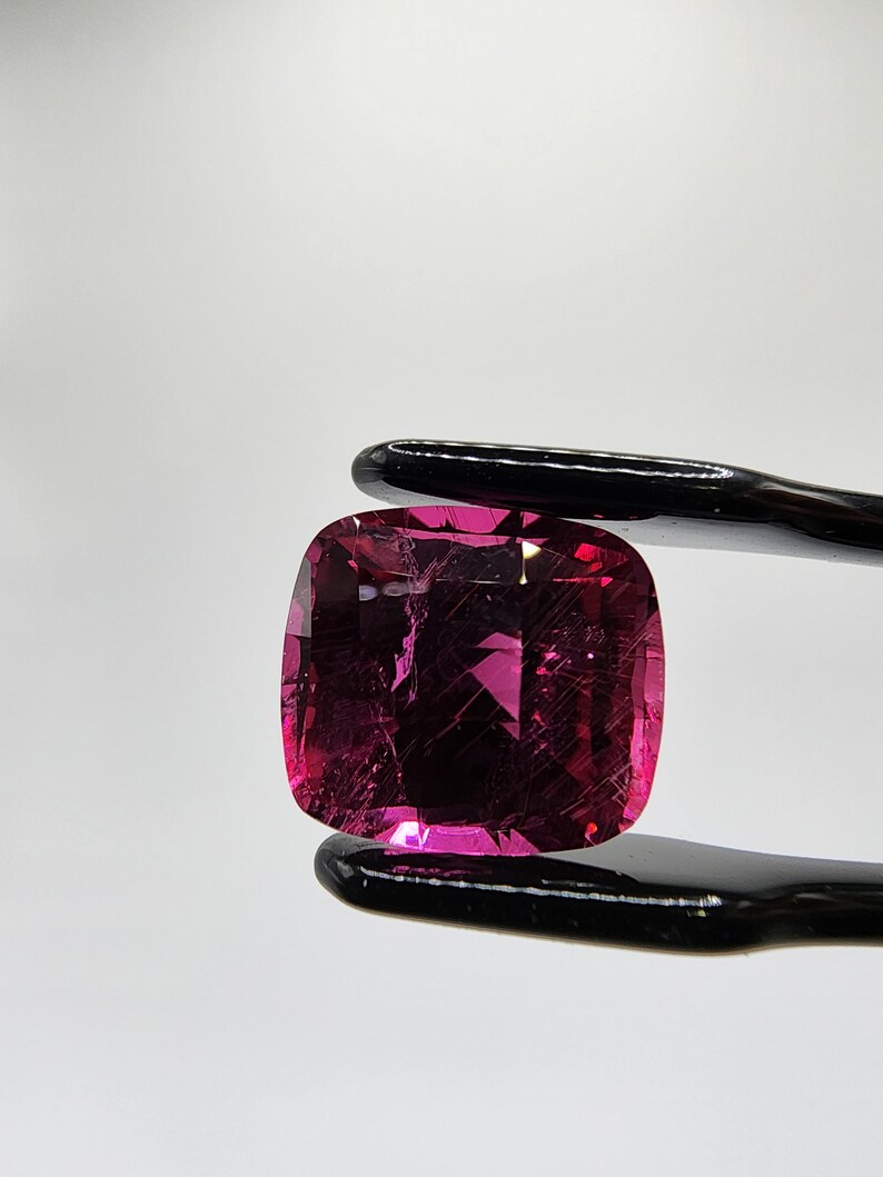 K&ouml;nnte beinhalten: Ein leuchtend rosa Edelstein mit Kissenform, gehalten von einer schwarzen Metallzange. Der Edelstein hat einen leichten Schimmer und einige Einschl&uuml;sse.