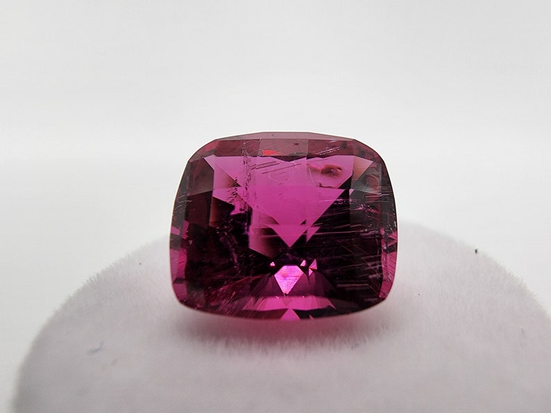 K&ouml;nnte beinhalten: Ein leuchtend rosa Edelstein mit einem Kissen-Schliff. Der Edelstein hat eine klare, facettierte Oberfl&auml;che und ist vor einem wei&szlig;en Hintergrund platziert.