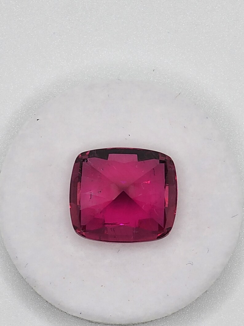 K&ouml;nnte beinhalten: Ein leuchtend rosa Edelstein mit einem Kissen-Schliff. Der Edelstein ist facettiert und hat einen hohen Glanz.