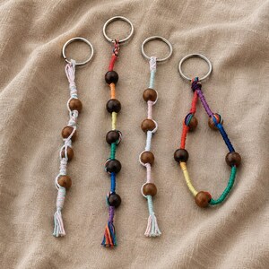 May include: Four keychains featuring metal rings and macrame cord in various colors including red, orange, yellow, green, blue, and pink. Each keychain incorporates smooth, dark brown wooden beads threaded onto the cord, with some designs ending in frayed tassels.