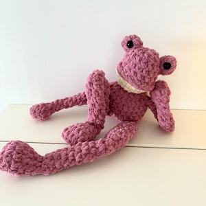 Pode incluir: Um brinquedo de pelúcia de sapo rosa feito à mão, com olhos pretos e boca branca. O sapo está deitado com as pernas longas estendidas. O texto "PATTERN RELEASE" está acima do sapo.