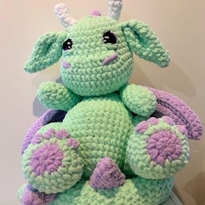 May include: A green crocheted dragon plush toy with purple accents. The dragon has white horns and large eyes.